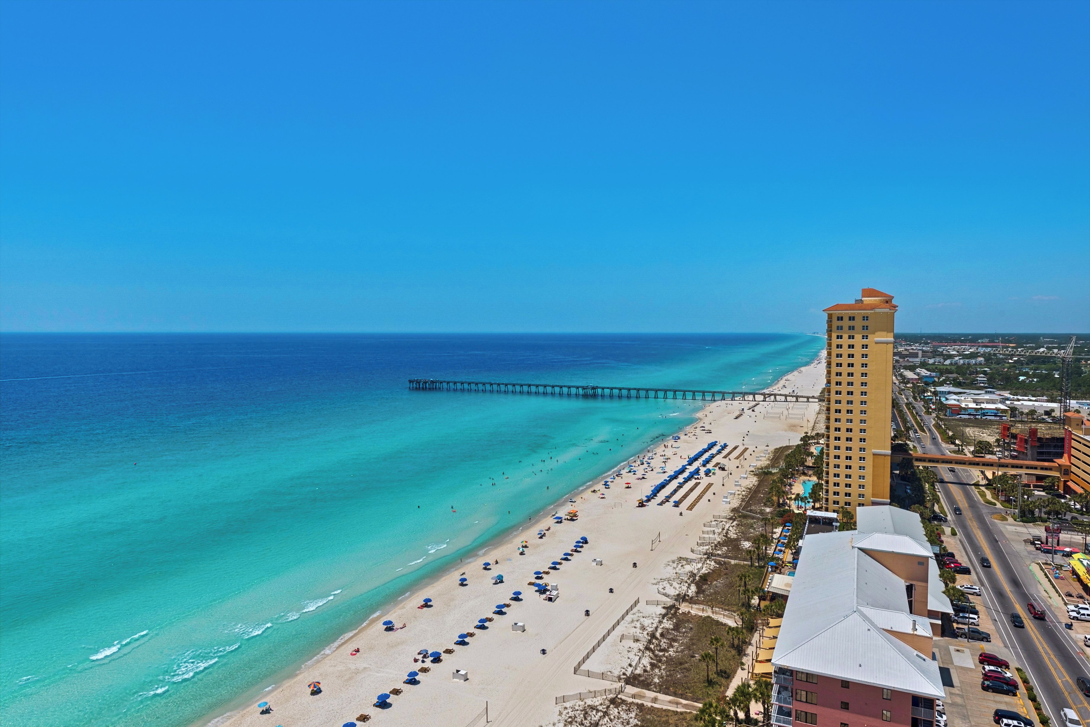 A beachside hotel with a long pier extending into the ocean, a sandy beach with umbrellas, and a clear blue sky.