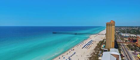 A beachside hotel with a long pier extending into the ocean, a sandy beach with umbrellas, and a clear blue sky.