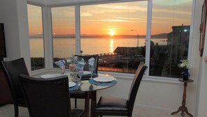 Een eethoek met een tafel gedekt voor twee personen, grote ramen met uitzicht op een zonsondergang en een decoratieve vaas op een bijzettafeltje.