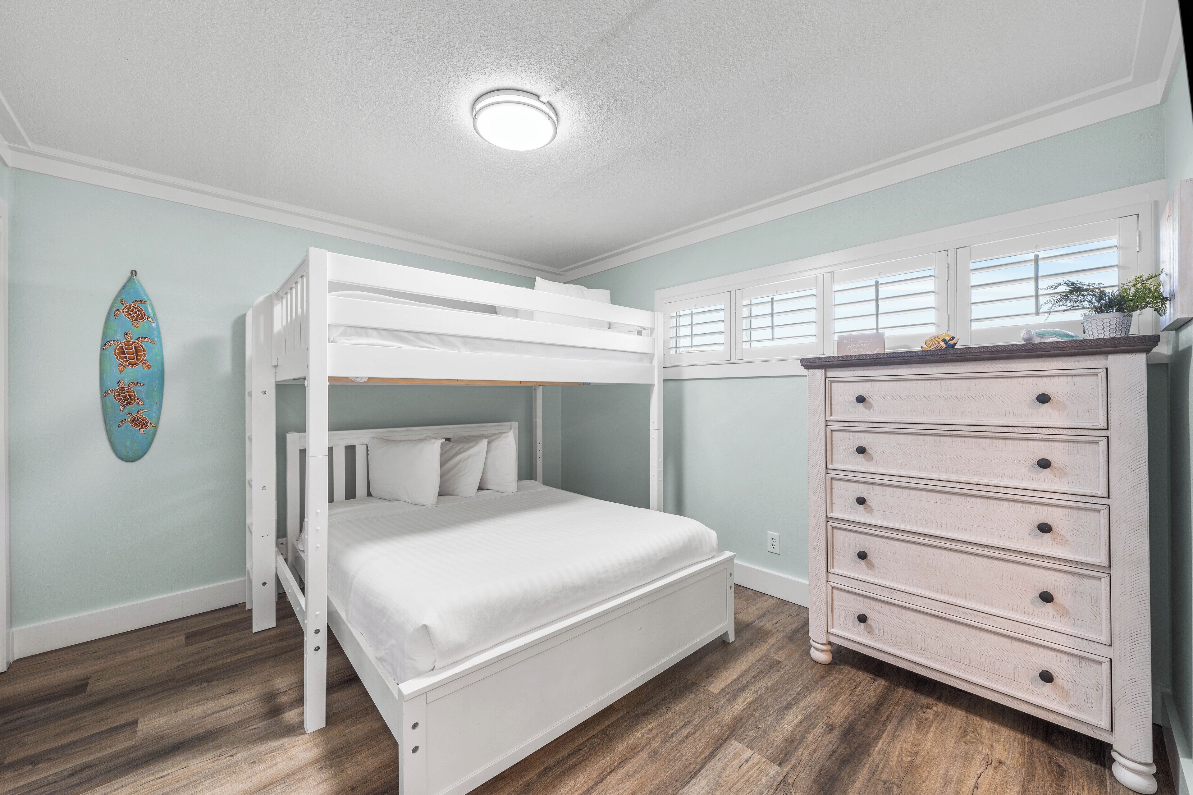 A bunk bed with a white frame and bedding, a dresser with drawers, a surfboard decoration, and a window with shutters.