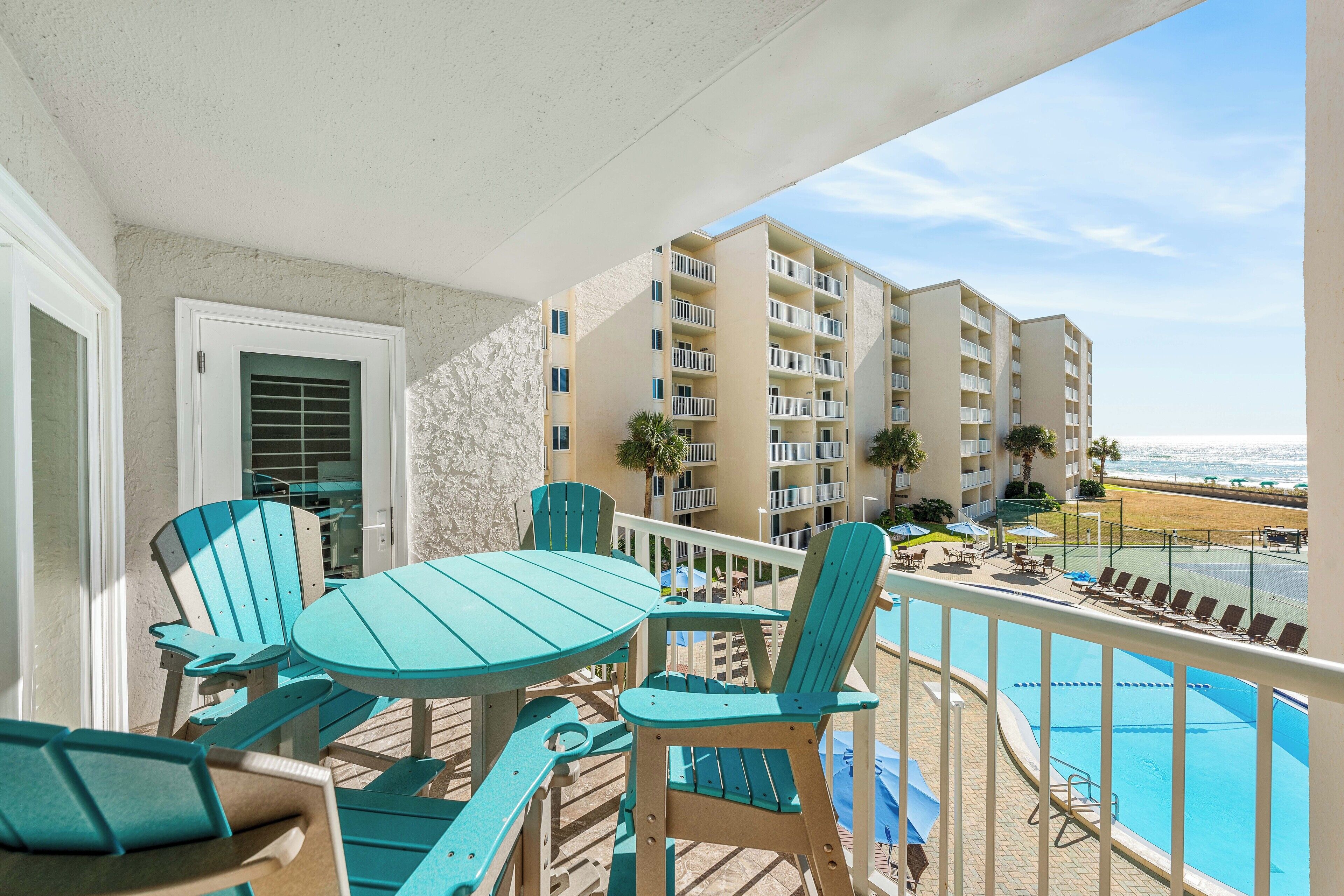 Un balcon avec des chaises turquoise et une table, donnant sur une piscine et sur la plage.