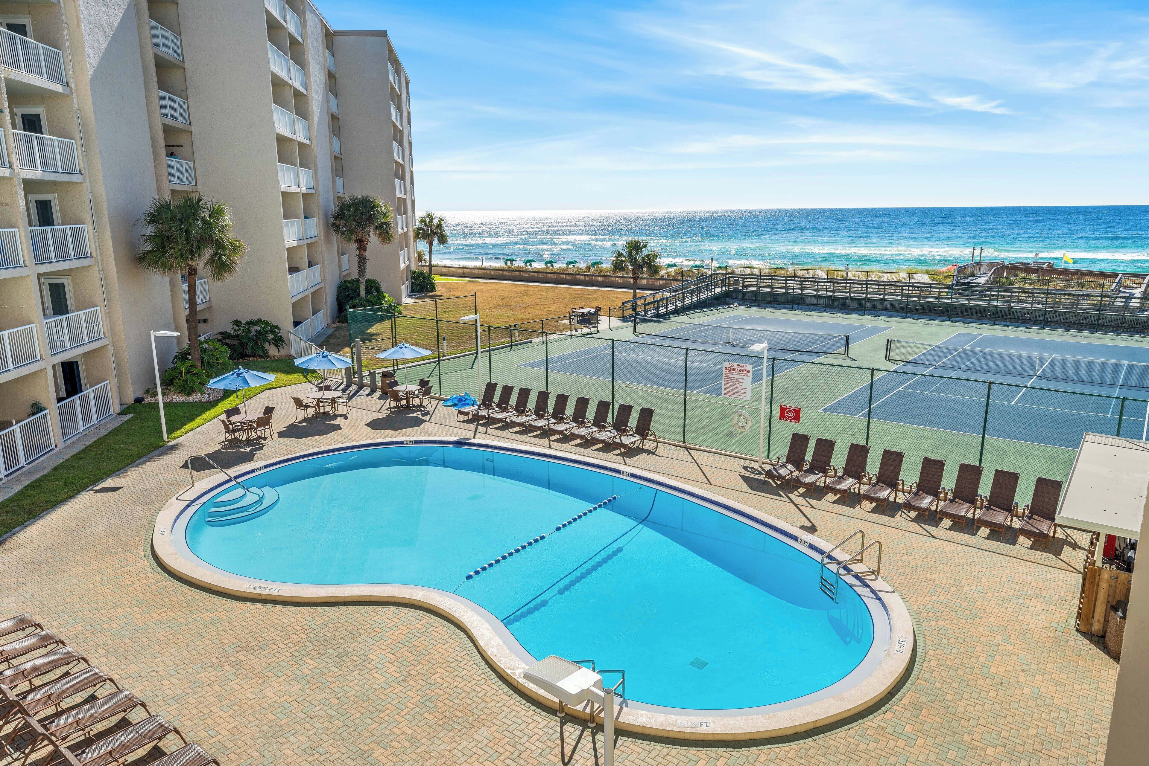 A swimming pool with a view of the ocean, surrounded by lounge chairs and a tennis court.