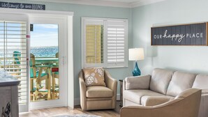 A living room with a beige armchair, a beige sofa, and a view of the ocean through a door with a sign that reads "Welcome to our HOME".