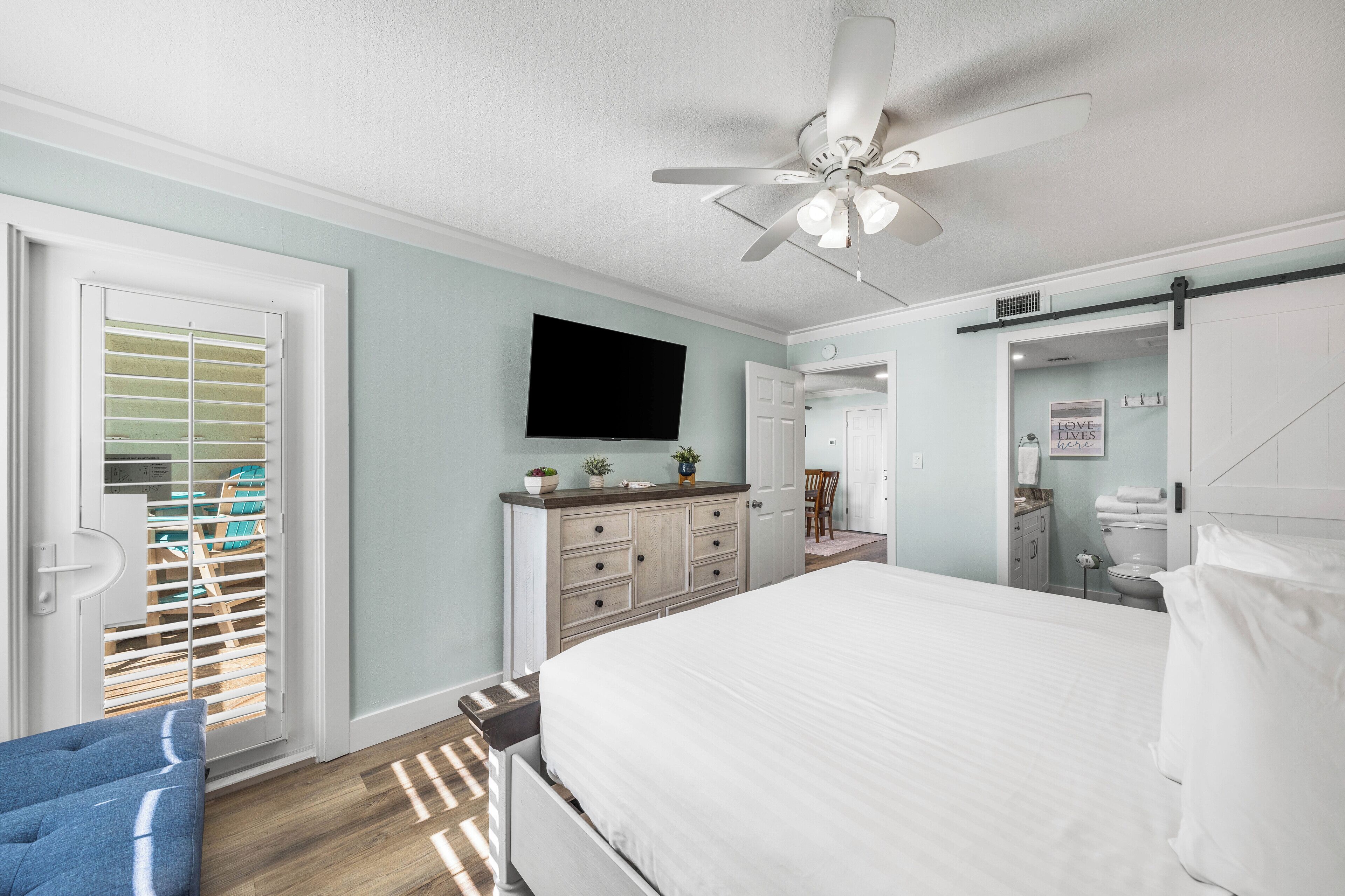 A bedroom with a bed, a dresser, a ceiling fan, a TV, and a sliding door leading to a balcony.