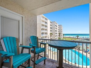 A balcony with two turquoise Adirondack chairs and a small table, overlooking a pool and a beach with buildings in the background.