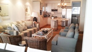 A modern living room with a sofa, armchairs, a coffee table, and a dining area with a kitchen in the background. - Waterfront Apartment C3 (Swakopmund)