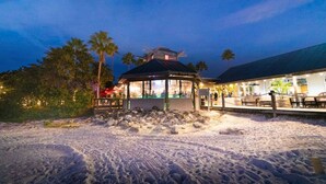 A beachside building with a covered seating area, palm trees, and a sandy foreground. - Salt Life Cottage U420 - Little Harbor Beach Resort Tampa Bay (Ruskin)