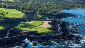 A coastal golf course with a rocky shoreline, lush greenery, and a mountainous backdrop.