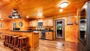 A wooden cabin kitchen with a bar area, wooden stools, a refrigerator, and a dishwasher. - American themed 9BR (sleeps 43) cabin in GBurg with tons of games and space! (GATLINBURG)