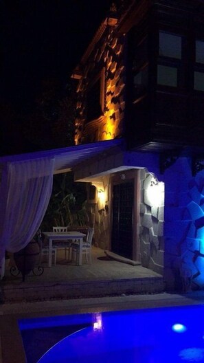 Vue nocturne d’un bâtiment doté d’un espace piscine, avec un mur en pierre et une terrasse couverte comprenant une table et des chaises.