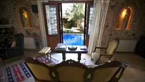 A cozy living room with a stone wall, a wooden sofa, a dining table with books, and a view of an outdoor swimming pool. - Mel'Dem Butik Otel (Cesme)
