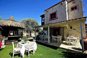 Una casa de piedra de dos pisos con un porche cubierto, mobiliario exterior y una estructura con techo de paja.