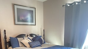 Una cama bien hecha con cabecera, un cuadro enmarcado en la pared y una ventana con cortinas.