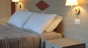 Een net opgemaakt bed met een beige deken en kussens, een klein nachtkastje met een lamp, en een wandlamp.