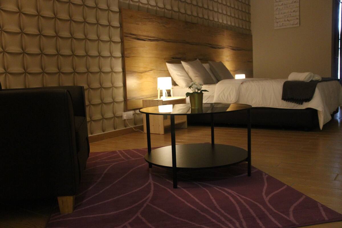 Habitación de hotel con cabecera capitonada, una mesa redonda, cama con sábanas blancas y una alfombra morada estampada.