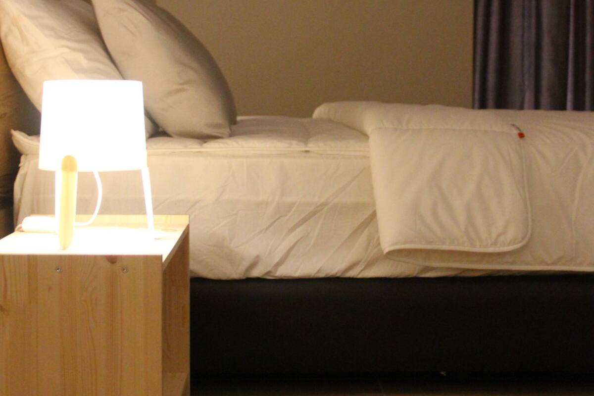 Una cama bien hecha con sábanas blancas, una lámpara de noche y una mesita de madera.