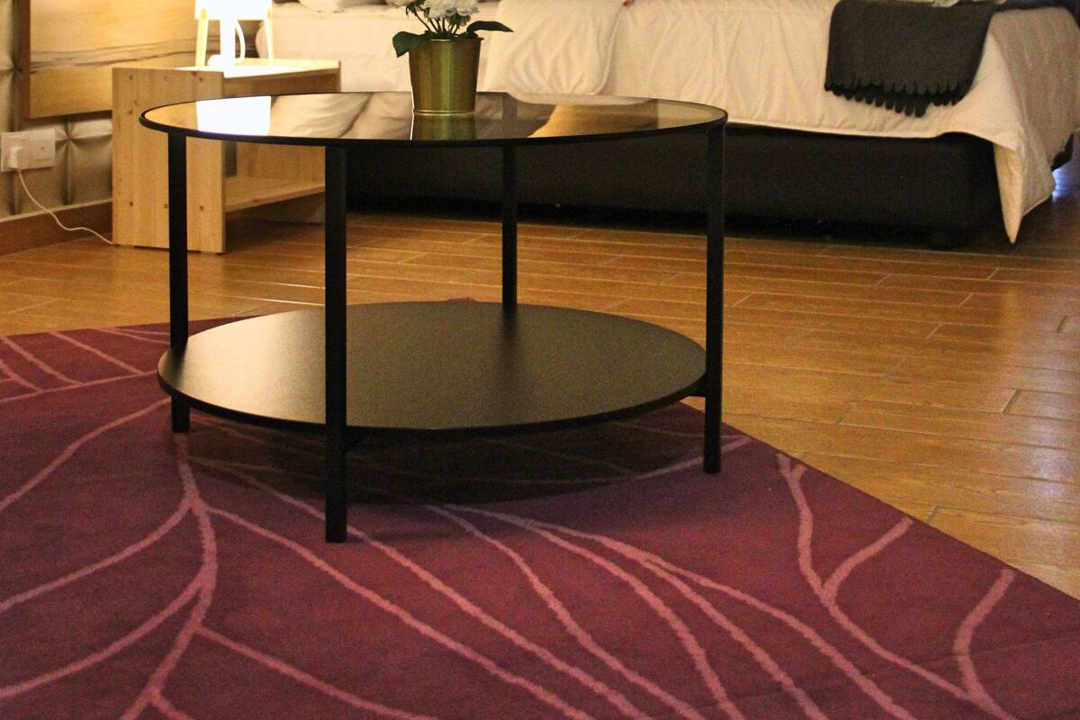 Una habitación de hotel con una cama, una mesita de noche con lámpara, una mesa redonda con un florero y una alfombra estampada.