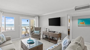 Een ruime woonkamer met een groot raam met uitzicht op het strand, een comfortabele zithoek, een houten salontafel en een flatscreen televisie aan de muur.