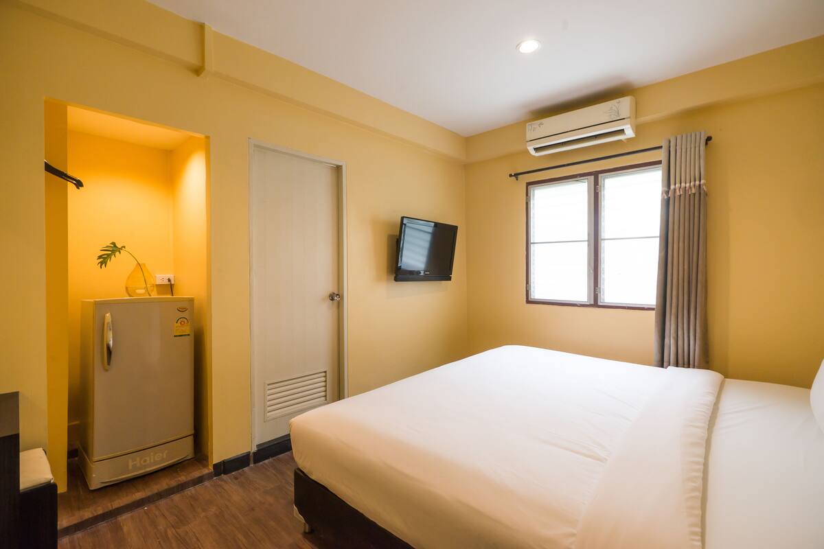 A hotel room with a bed, a television mounted on the wall, an air conditioning unit, and a door leading to a hallway.