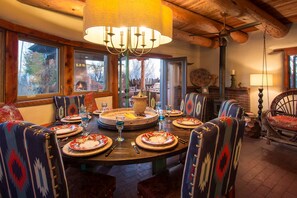 Un coin repas avec une table ronde dressée pour six personnes, entourée de chaises à motifs, d’un lustre et d’une cheminée en arrière-plan.
