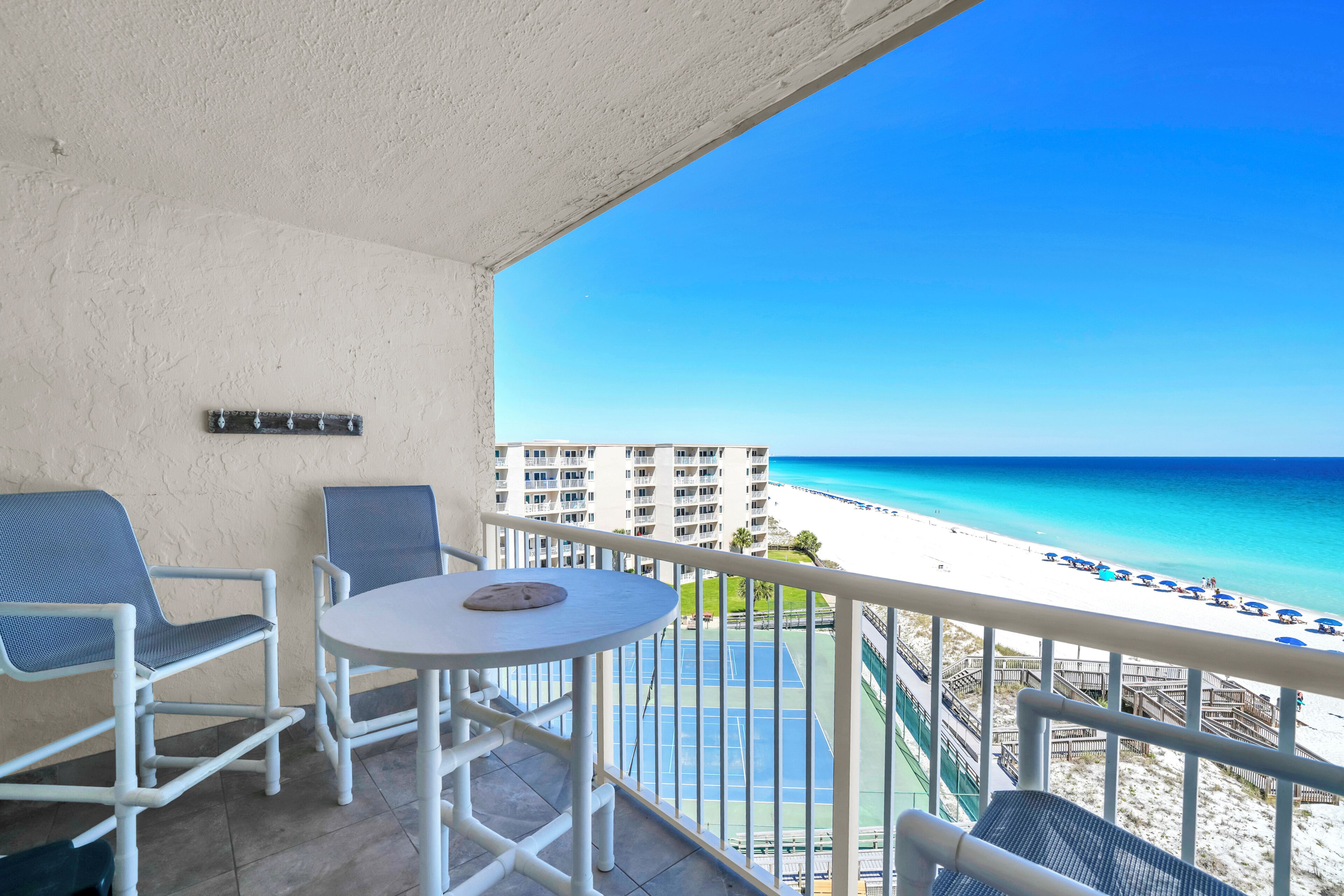 A balcony with a white table and two chairs, overlooking a beach with a clear blue sky and a view of a hotel building in the distance.