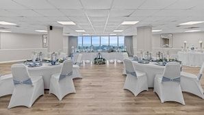 Een ruime, goed verlichte congreszaal met uitzicht op de oceaan door grote ramen, tafels met witte tafellakens en blauwe linten, en witte stoelen met blauwe linten.
