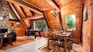 A cozy log cabin interior with a dining area, wooden beams, a fireplace, and a large window. - A-Frame cabin- 3BR, pet friendly, hot tub, located between GBurg & Pigeon Forge! (Sevierville)