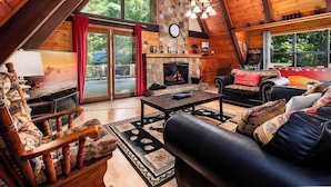 A cozy cabin interior with a stone fireplace, wooden furniture, and large windows offering a view of the outdoors. - A-Frame cabin- 3BR, pet friendly, hot tub, located between GBurg & Pigeon Forge! (Sevierville)