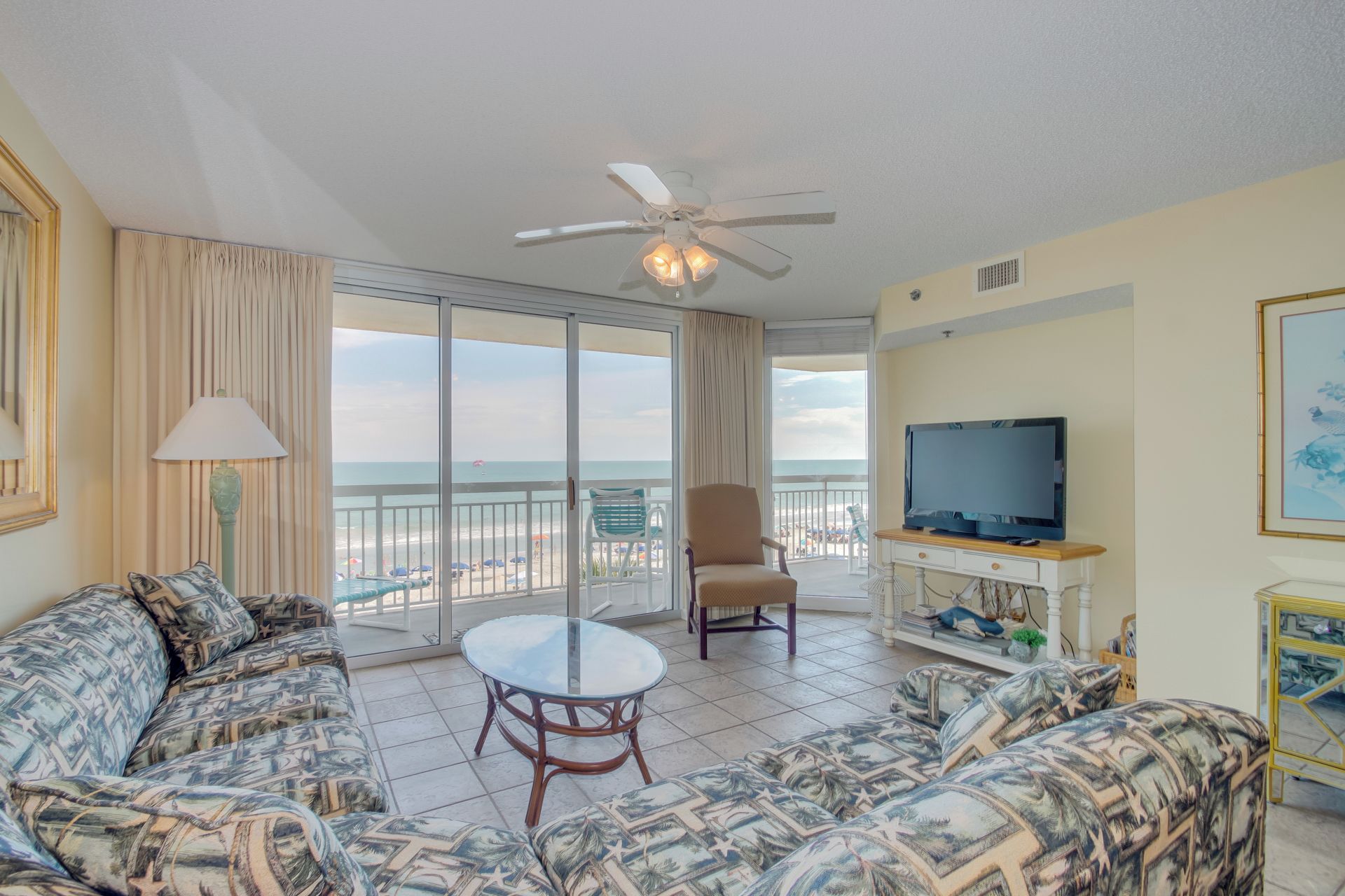 A living room with a sofa, armchair, and TV, overlooking a beach.