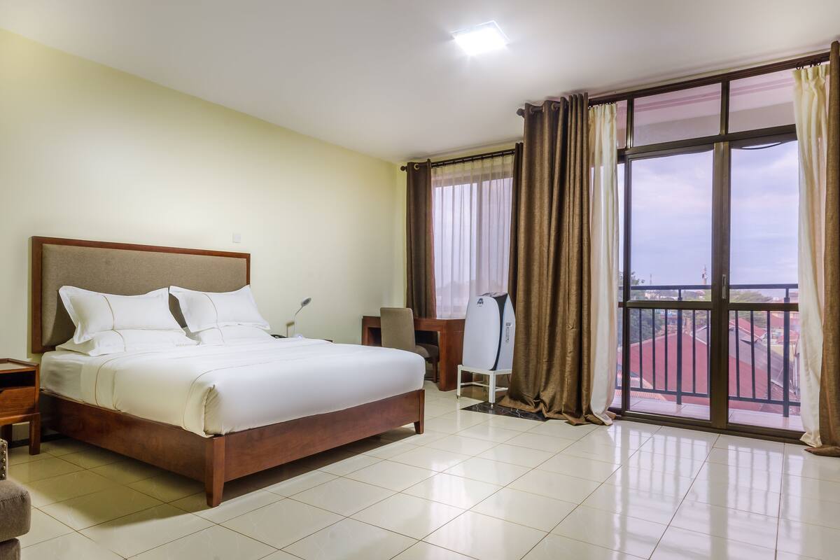 A hotel room with a large bed, a desk with a chair, a window with curtains, and a view of buildings.