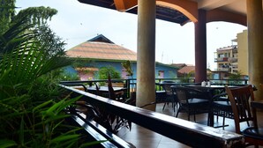 An outdoor patio area with tables and chairs, surrounded by greenery and buildings in the background.
