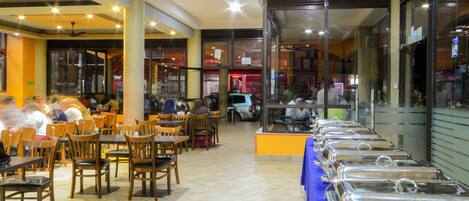 Una cafetería con varias mesas y sillas, una barra de buffet con recipientes de comida y un ventilador de techo.