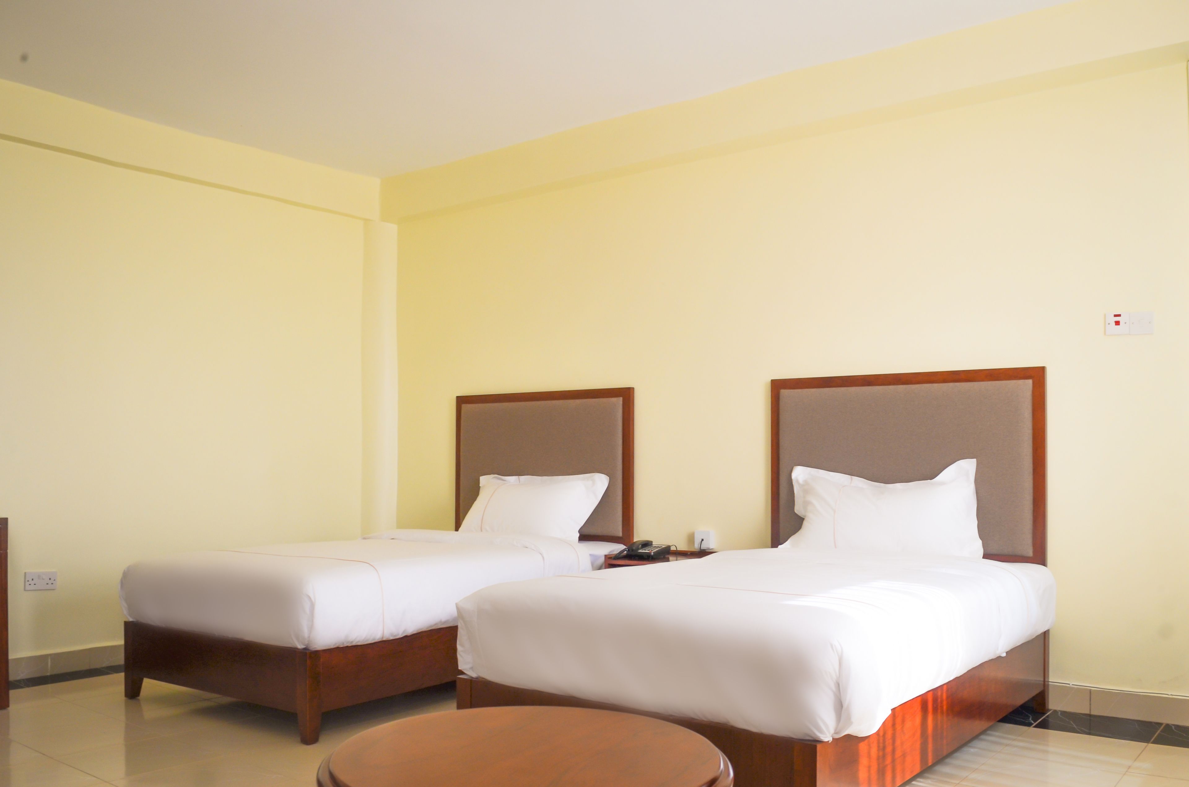 Habitación de hotel con dos camas individuales, un cabecero de madera, una mesita de noche con un teléfono y un taburete redondo de madera.