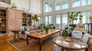 A spacious living room with a high ceiling, large windows, and a variety of plants and decorative items. - Softwind |fall openings. community pool. gulf front (Dauphin Island)