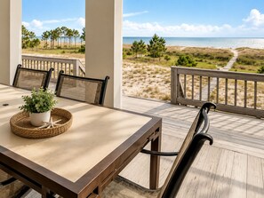 Une terrasse en bois avec une table et des chaises, donnant sur une plage bordée de dunes de sable et d’arbres.