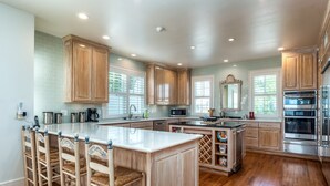 A spacious kitchen with wooden cabinets, a large island with bar stools, stainless steel appliances, and a view of the outdoors through windows. - Softwind |fall openings. community pool. gulf front (Dauphin Island)