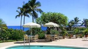 A pool area with lounge chairs and umbrellas, surrounded by palm trees and a view of the ocean.