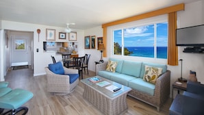 A living room with a sofa, chairs, a coffee table, and a TV. There is a window with a view of the ocean.