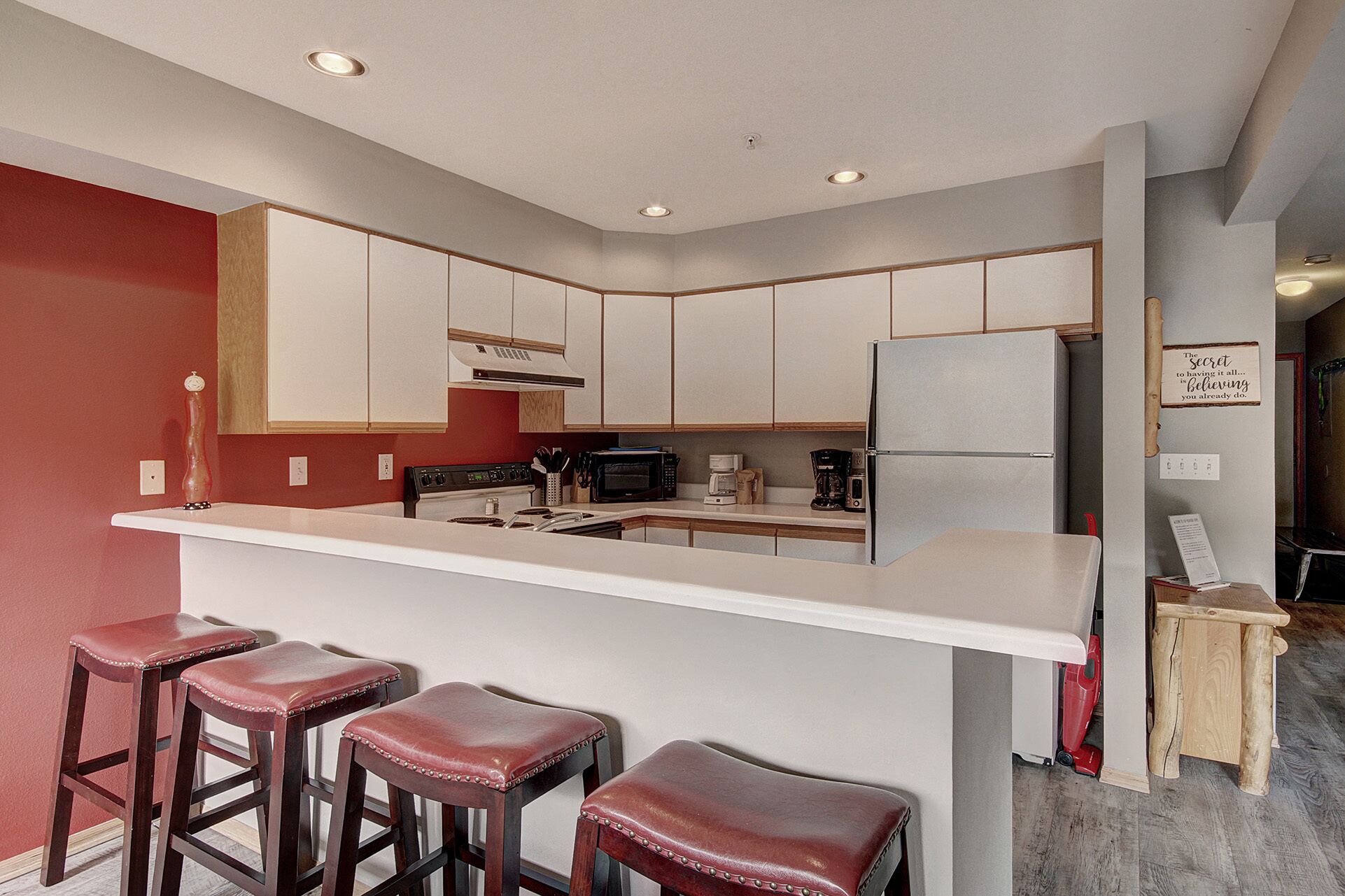 Una cocina moderna con armarios blancos, un refrigerador de acero inoxidable, un microondas y una cafetera. Hay tres taburetes de madera con cojines rojos.