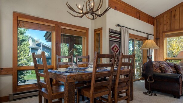 A dining area with a wooden table and chairs, large windows, and a chandelier. - JHRL - Nez Perce B6 - Great location near pool and hot tub access (Teton Village)
