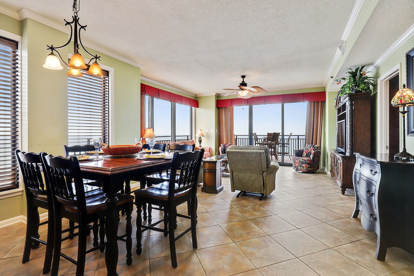 A spacious dining area with a wooden table and chairs, a ceiling fan, and large windows with red curtains.