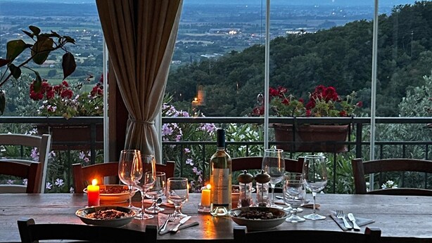 Mesa de comedor dispuesta con velas, copas y cubiertos, con vistas a un paisaje pintoresco con montañas y vegetación.