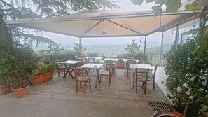 Un patio al aire libre con piso de piedra, mesas y sillas de madera, rodeado de plantas en macetas y vistas panorámicas.