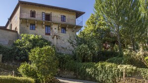 A two-story stone building with a balcony, surrounded by greenery and a stone wall. - Torre Lombarda (Allariz)
