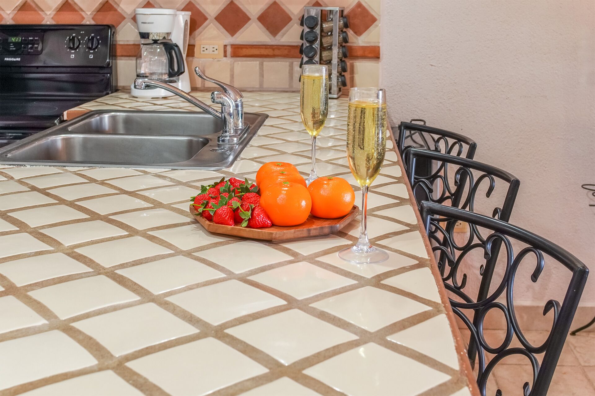 A kitchen with a tiled backsplash, a stainless steel sink, a coffee maker, a wine rack, and a fruit bowl with oranges and strawberries.