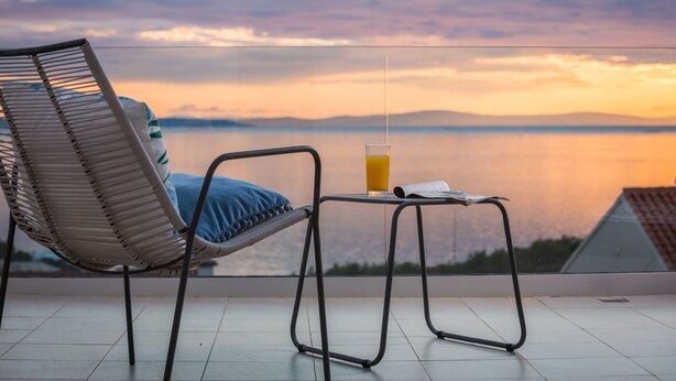 Un balcón con una silla de mimbre, una pequeña mesa con un vaso de zumo de naranja y vistas a un cuerpo de agua tranquilo y montañas lejanas al atardecer.