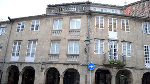 Ein historisches Steinhaus mit zahlreichen Fenstern, einem blauen Hinweisschild und einem Blumenkasten am Balkon.