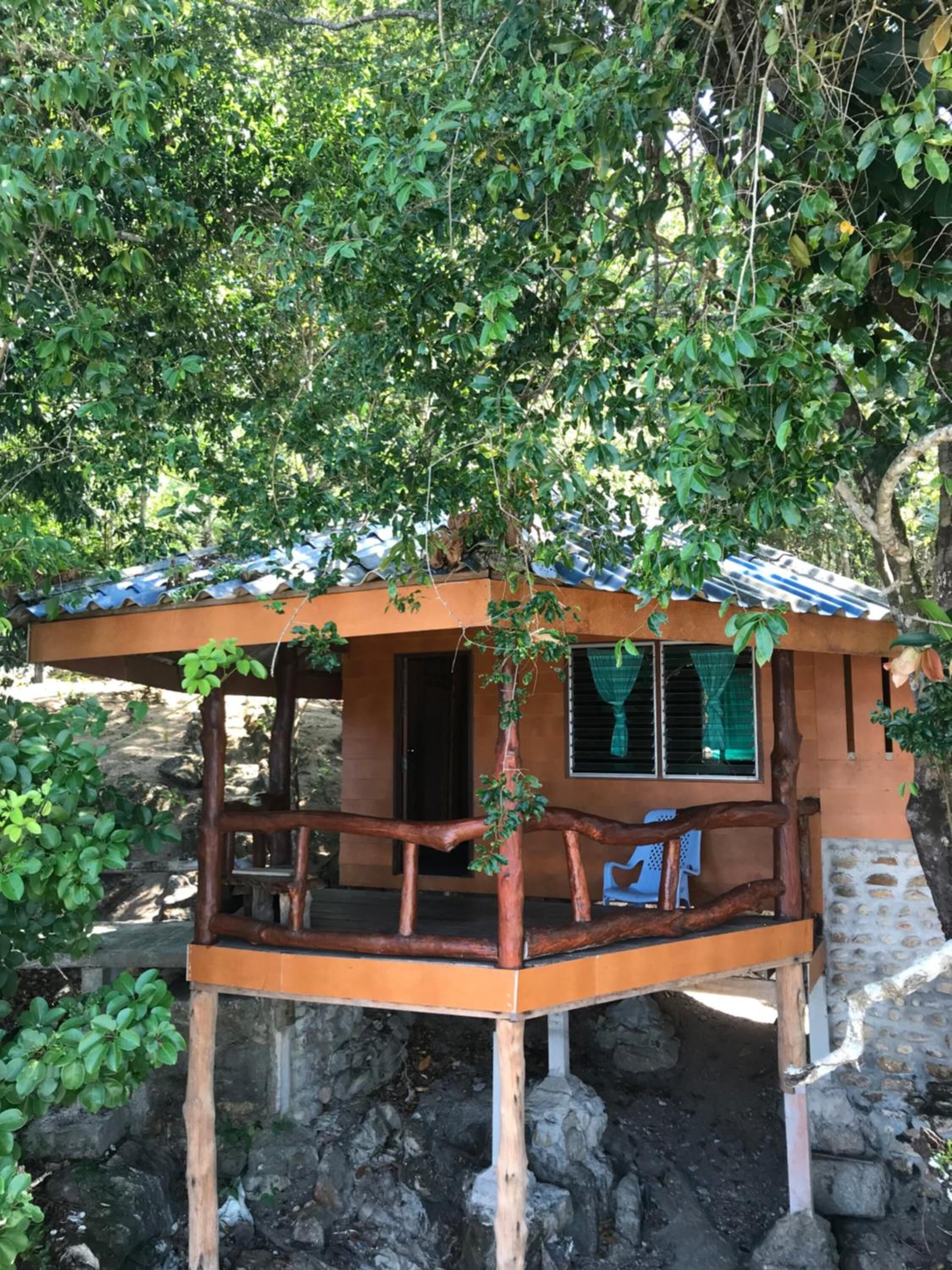 Una cabaña de madera rústica con porche, rodeada de abundante vegetación y una base rocosa.