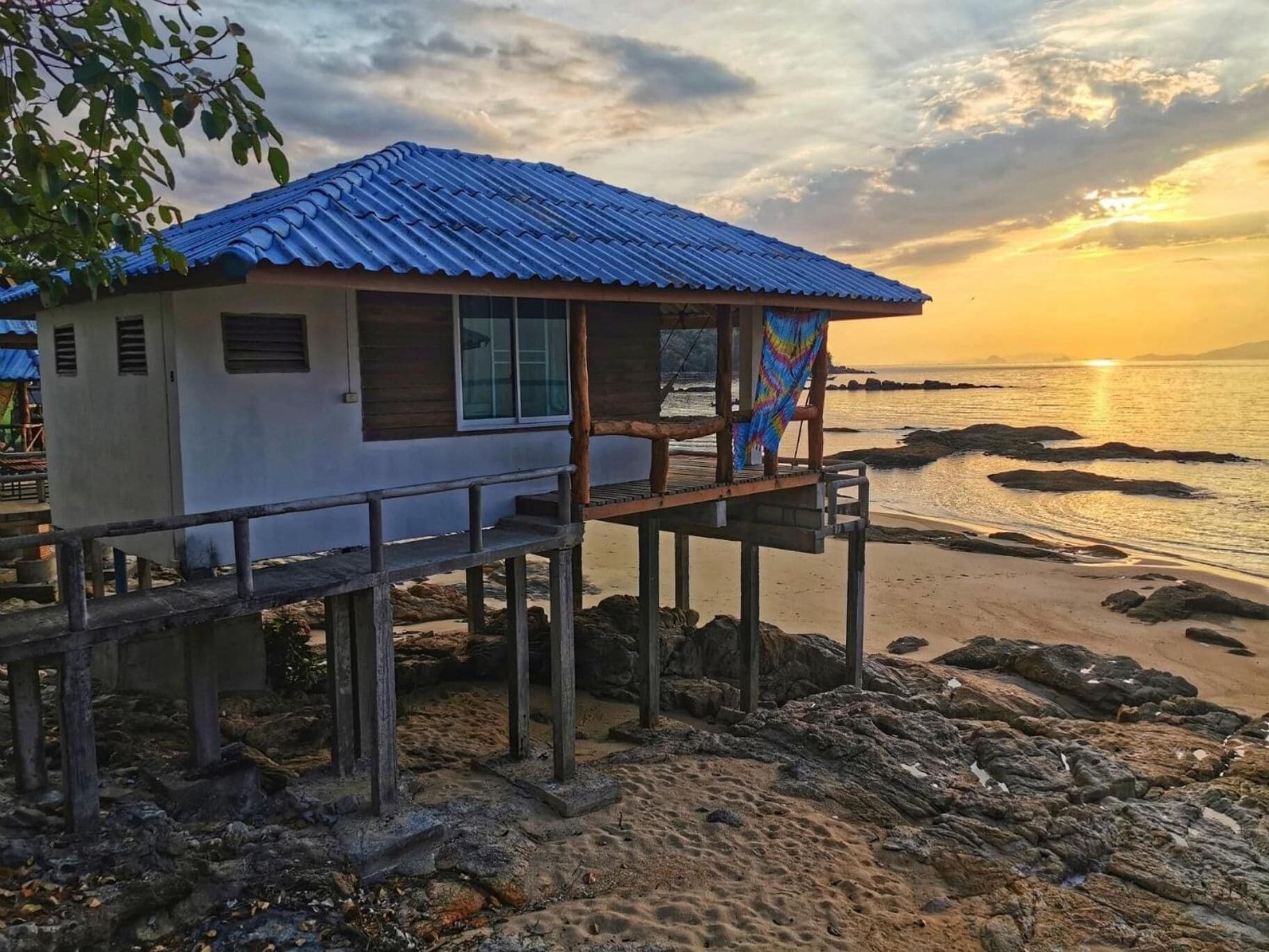 A small building with a blue roof on a wooden pier, overlooking a beach with rocks and a sunset.