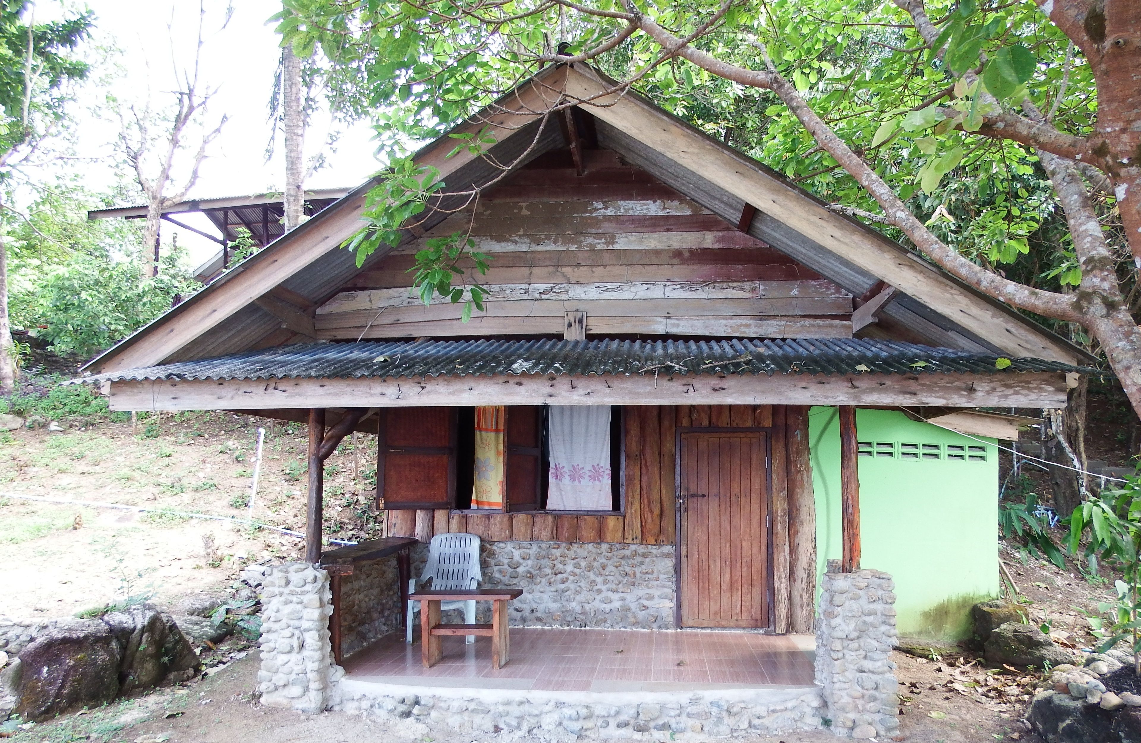 A rustic wooden cabin with a sloped roof, a porch with a single chair, and a stone wall foundation.